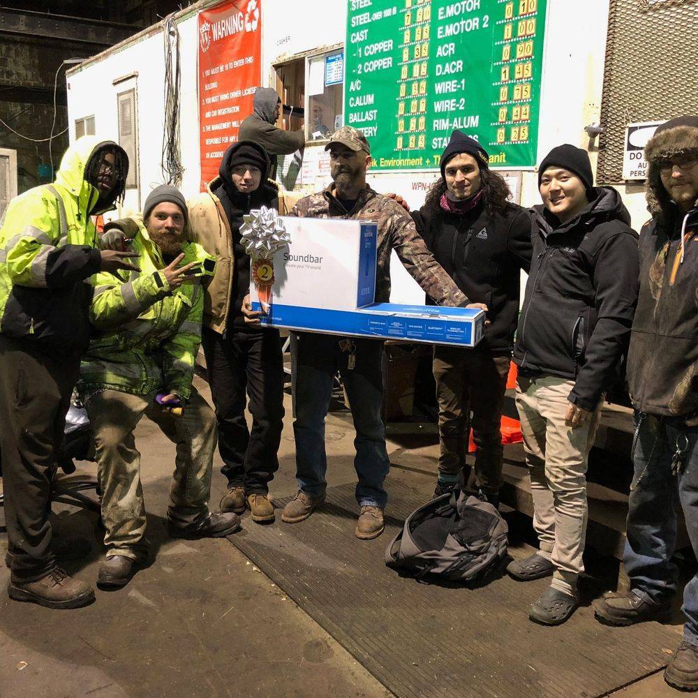 Seven people standing in the warehouse holding a boxed television, in front of a price board.